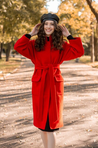 暖かい赤いコートの秋の流行のファッション、ストリートスタイル、ベレー帽の帽子をかぶって公園を歩いている巻き毛の幸せなかわいい魅力的なスタイリッシュな笑顔の女性