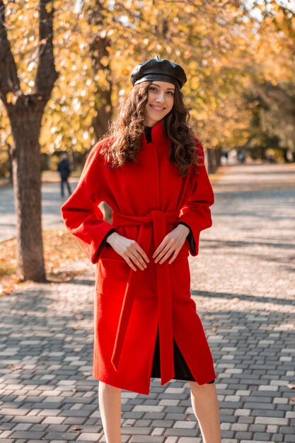 暖かい赤いコートの秋の流行のファッション、ストリートスタイル、ベレー帽の帽子をかぶって公園を歩いている巻き毛の幸せなかわいい魅力的なスタイリッシュな笑顔の女性