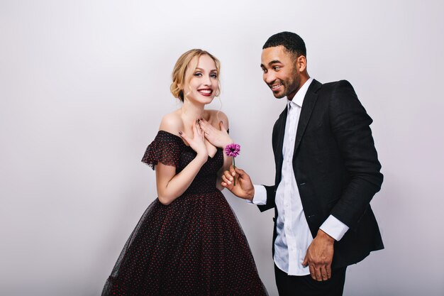 Happy cute couple in-love celebrating Valentines day. Attractive young blonde woman in luxury dress, handsome man in tuxedo, giving flower, smiling, positive emotions.