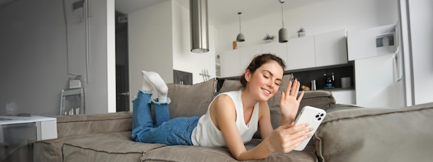 Бесплатное фото Счастливая милая модель лежит на диване и махает рукой по смартфону, говоря 