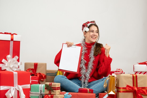 happy cute girl with santa hat holding document sitting around presents on white