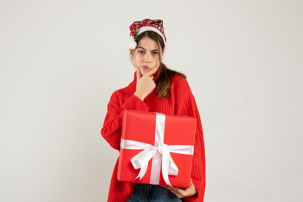 happy cute girl with santa hat holding heavy gift putting hand on her chin standing on white