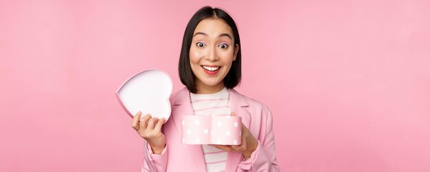 Happy cute korean girl in suit opens up heart shaped box with romantic gift on white day holiday standing in suit over pink background