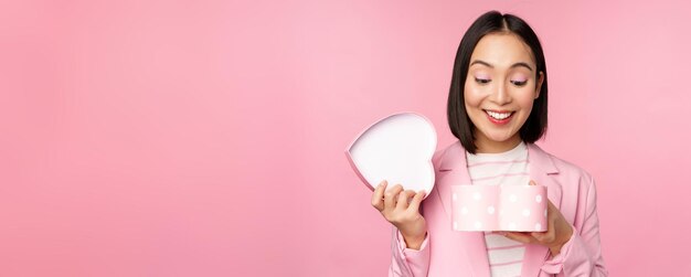 Happy cute korean girl in suit opens up heart shaped box with romantic gift on white day holiday standing in suit over pink background