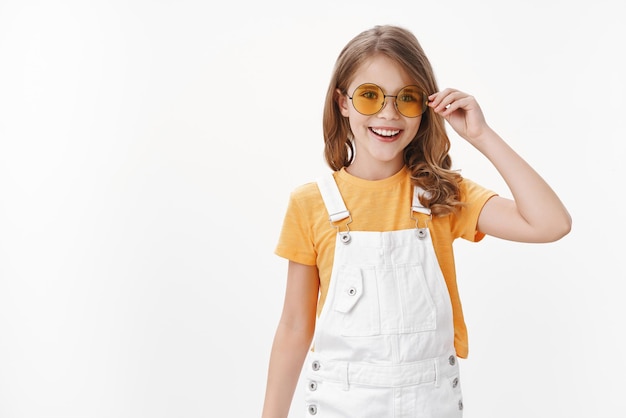 Free photo happy cute little fashionable girl, daughter wearing yellow round sunglasses, t-shirt and dungarees