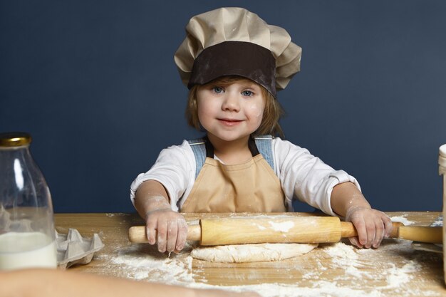 Happy cute little girl flattening pastry using rolling pin while helping mother cooking pie for dinner. Sweet female child with blue eyes making cookies in the kitchen, looking and smiling at camera