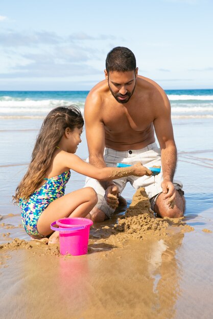 Happy cute little girl and her dad building sandcastle on beach, sitting on wet sand, enjoying vacation