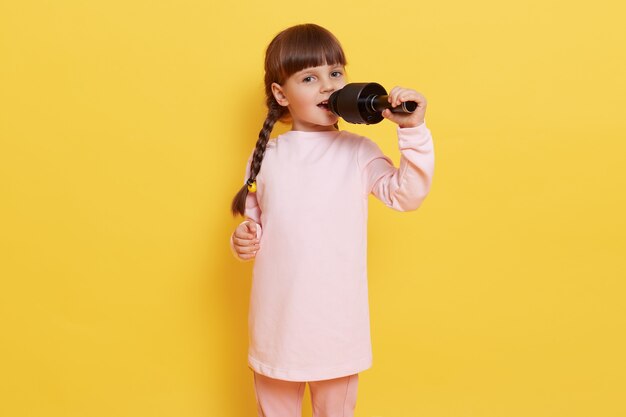 Happy cute little girl singing song on microphone while posing isolated over yellow background, dark haired female chile with pigtails sings in karaoke, looking at camera with excited and happy look.