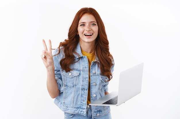 Happy cute redhead female freelancer in denim jacket easily accomplish goal using laptop, show peace victory, goodwill sign, smiling joyfully, working on remote, prepare project, white wall