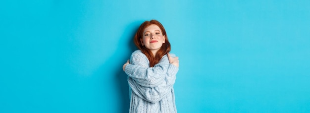 Free photo happy cute redhead girl hugging herself wearing comfortable and warm sweater smiling at camera stand