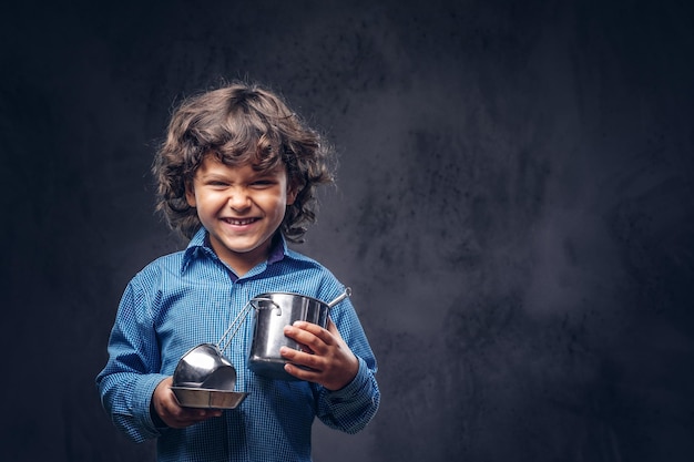 Free photo happy cute schoolboy with brown curly hair dressed in a blue shirt holds a cookware at a studio. isolated on the dark textured background.