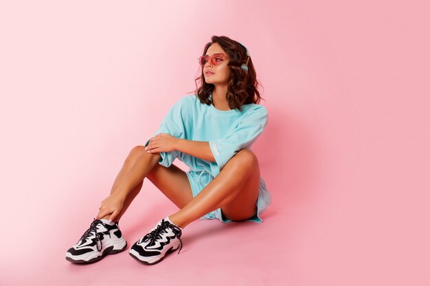 Happy cute woman listening music by earphones, wearing blue outfit, stylish trainers sitting on pink