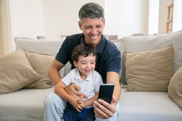 Happy dad and adorable little son having fun together, using phone for video chat while sitting on couch at home