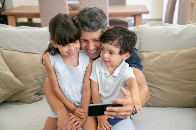 Счастливый папа держит двух детей на коленях, делает селфи или использует телефон для видеозвонка
