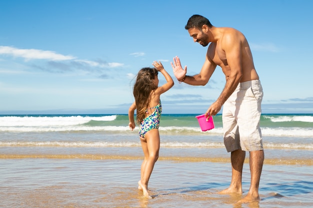 Happy dad and little daughter picking shells with bucket on beach together, giving high five