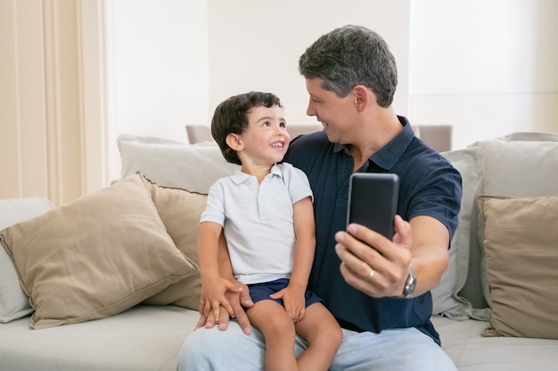 Happy dad and little son enjoying time together, sitting on couch at home, chatting, laughing and taking selfie.