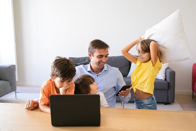 Happy dad using smartphone, sitting at table and kids playing with him. Caucasian father working at home, using laptop and watching children. Fatherhood, childhood and digital technology concept