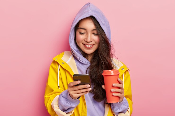 無料Photo 幸せな黒髪の少女は、パーカーとレインコートを着て、携帯電話を持って、社会のネットワークでフィードをスクロールし、コーヒーやお茶を飲みます