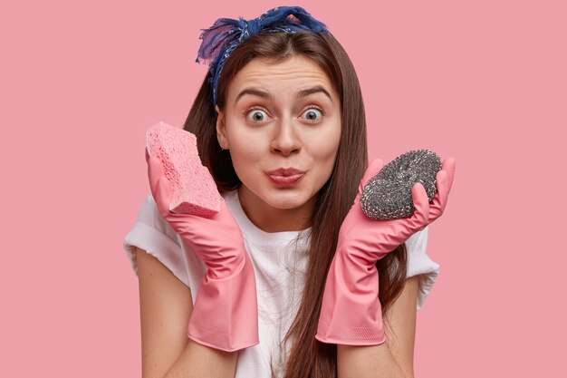 Happy dark haired woman cares of sanitary and hygiene, wears rubber gloves, holds two mops in hands, prepares to clean filthy bathroom, wears white t shirt