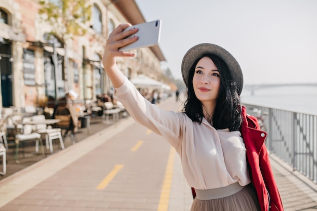 Happy dark-haired woman in romantic outfit making selfie near street cafe
