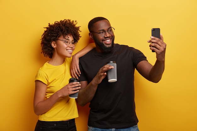 Happy dark skinned couple in love have fun during coffee break, make selfie portrait on modern cell phone, wear round spectacles