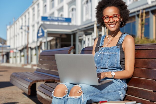 Happy dark skinned female designer watches tutorial about creative ideas, keeps portable laptop computer on knees, listens online news with earphones, wears spectacles and denim overalls poses outdoor