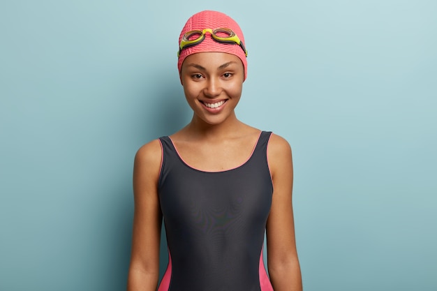 Happy dark skinned female going to swim, wears pink swimcap and goggles