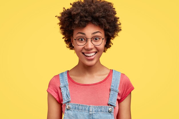 アフロの髪型をした幸せな暗い肌の女性は、表情を喜ばせ、白い完璧な歯を見せ、彼氏とのデートの後、黄色い壁に隔離されて気分が良い。感情の概念