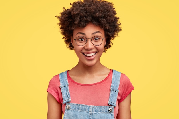 Happy dark skinned female with Afro hairstyle, has pleased expression, shows white perfect teeth, being in good mood after date with boyfriend, isolated over yellow wall. Emotions concept