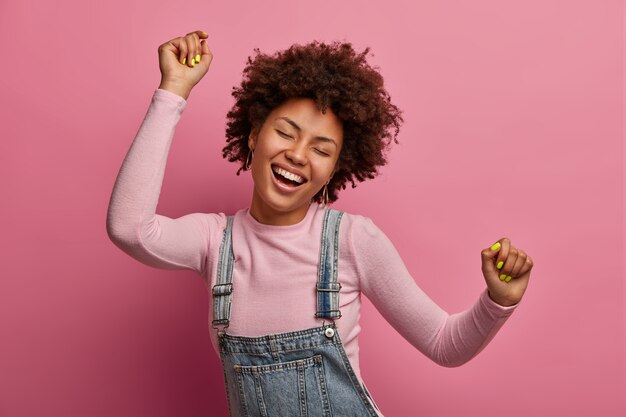Happy dark skinned girl enjoys every moment of life, dances and moves, raises arms and clenches fists, closes eyes, has good mood, wears denim sarafan and turtleneck, isolated on pink wall