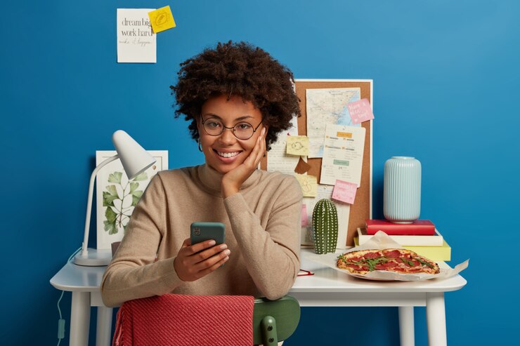 Free Photo happy dark skinned girl enjoys free high speed internet, uses mobile phone for texting, sits against workplace