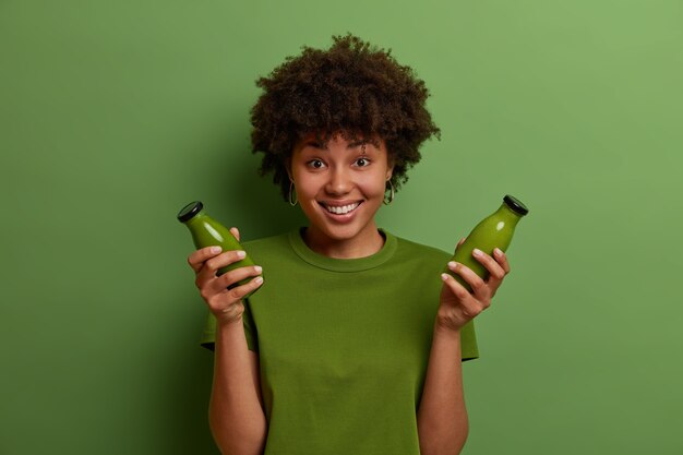 幸せな暗い肌の女の子は、生の緑のデトックス野菜のスムージーとガラス瓶を持って、健康的なライフスタイルを導き、菜食主義の食事を続け、さわやかで嬉しいと感じます。モノクロームショット。人と健康
