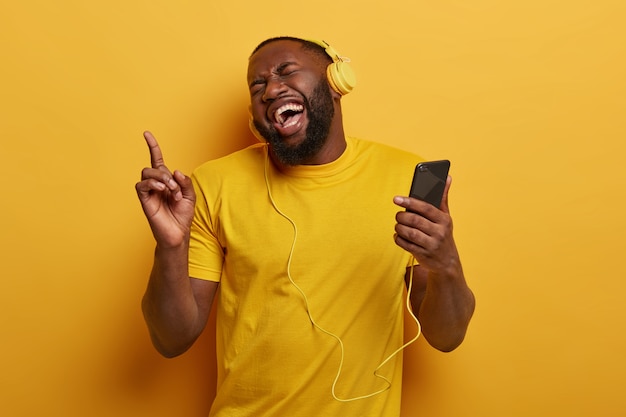 Happy dark skinned hipster guy with thick beard, laughs joyfully, points index finger above, holds modern cell phone, listens music in headphones