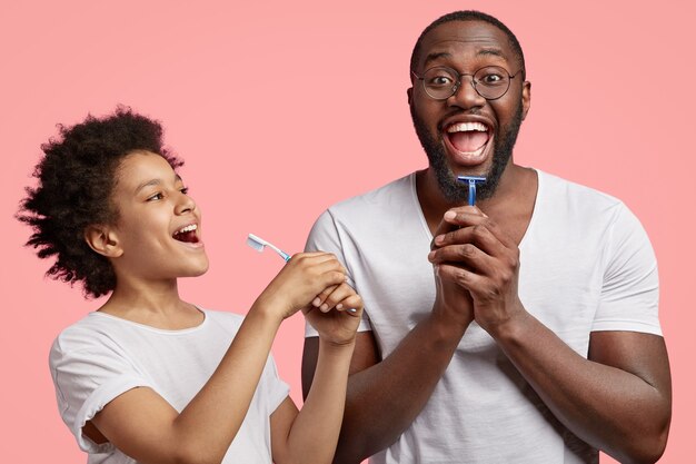 Happy dark skinned joyful dad and son hold toothbrsuh and shaver, have morning routine together