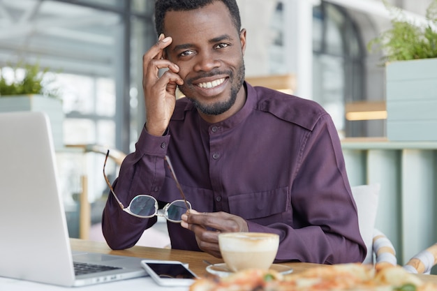 Felice libero professionista maschio dalla pelle scura utilizza moderni gadget elettronici per il lavoro a distanza, si siede contro l'interno accogliente della caffetteria, beve caffè espresso aromatico