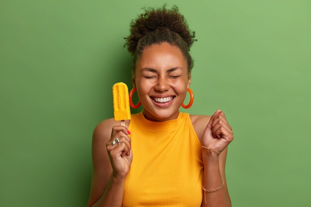 Happy dark skinned millennial girl eats yellow ice cream of mango flavor, clenches fist with anticipation of something awesome happen, has toothy smile, wears bright summer clothes, isolated on green