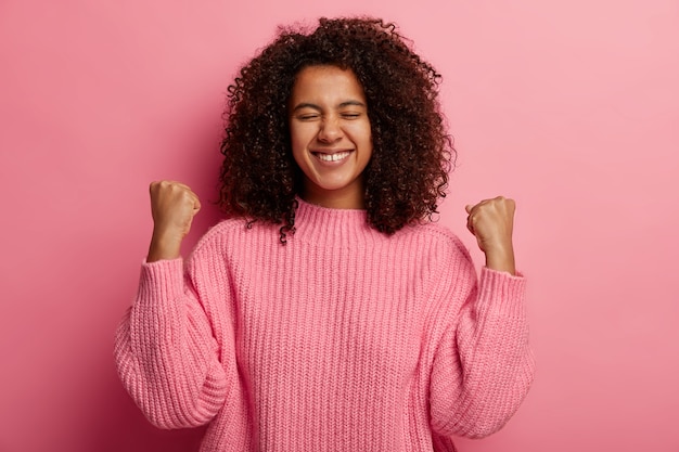 Foto gratuita felice adolescente dalla pelle scura solleva i pugni chiusi, celebra la vittoria e il successo, ha ottenuto il desiderio desiderabile, sorride ampiamente, vestito con un maglione lavorato a maglia, isolato sul muro rosa.