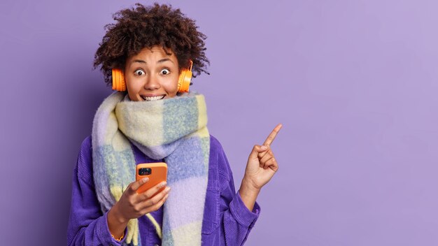 행복한 어두운 피부를 가진 여자는 따뜻한 겨울 스카프에 싸인 곱슬 머리가 있습니다. 온라인 커뮤니케이션을 위해 휴대 전화를 보유하고 있으며 오른쪽에 놀라운 제안 포인트를보고 놀란 귀에 헤드폰을 착용합니다.