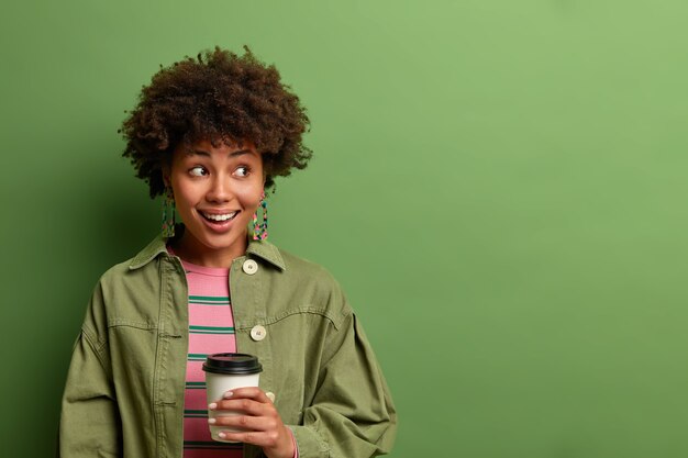 happy dark skinned woman holds disposable cup of hot drink, looks aside and smiles gladfully, takes coffee break, enjoys morning caffeine beverage, isolated on green wall, copy space