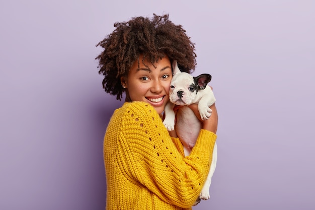 Free photo happy dark skinned woman holds tenderly her small bulldog puppy, makes portrait in studio