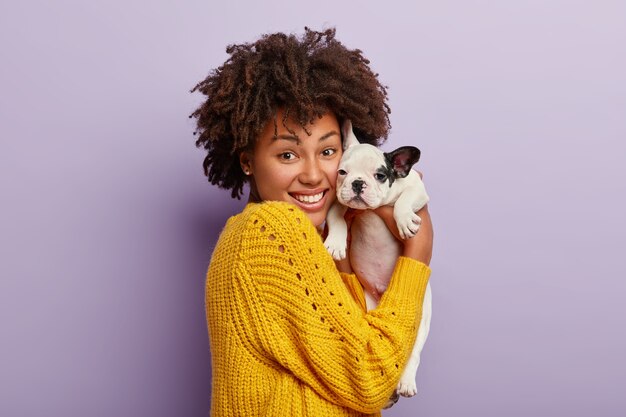 Happy dark skinned woman holds tenderly her small bulldog puppy, makes portrait in studio