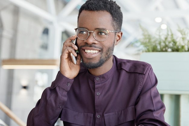 Happy dark skinned young male with positive expression, has broad smile, dressed in formal clothing, has telephone conversation with business partner