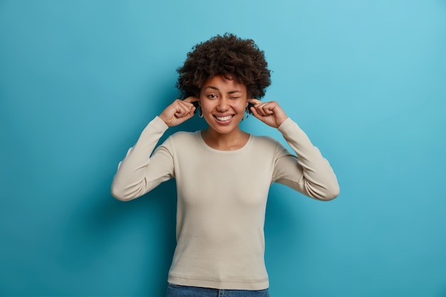 Happy dark skinned young woman ignores loud music and plugs ears with fingers