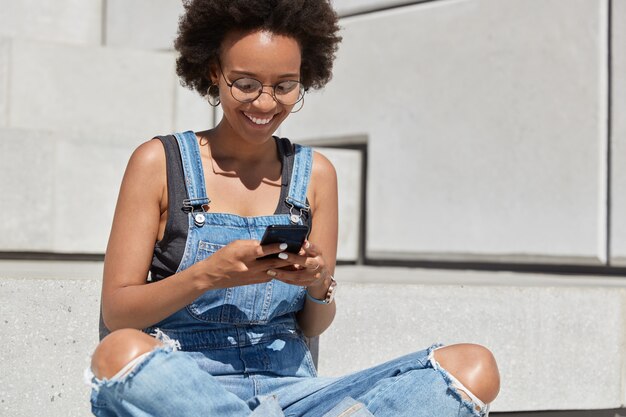 巻き毛のヘアカットで幸せな暗い肌の若者、楽しいテキストメッセージを読み、面白いビデオを見て、フィードバックを入力し、外の階段で蓮華座に座って、デニムのぼろぼろのダンガリーを着て、オンラインでサーフィン