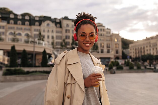 Happy darkskinned woman holds orange juice glass Charming lady in beige trench coat listens to music in red headphones outside