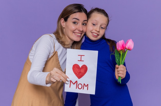Happy daughter giving greeting card and tulips flowers for her surprised and smiling mother celebrating mother's day standing over purple wall