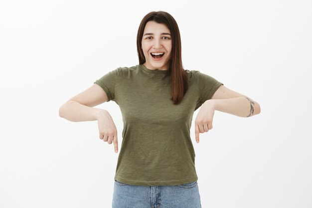 Happy and delighted bright optimistic female with brown hair and eyes in olive t-shirt smiling broadly and pointing down
