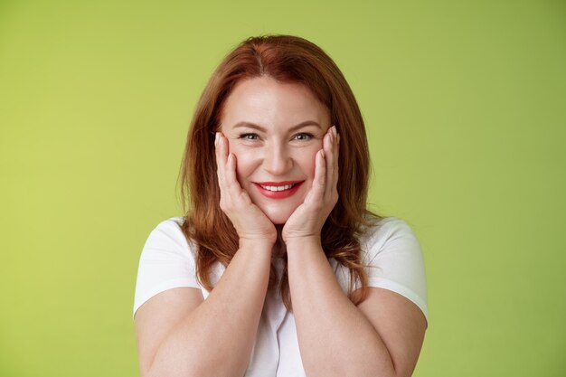 Happy delighted cheerful lucky redhead middleaged caucasian woman blushing joyfully receive touching cute gift touch cheeks pleased delighted smiling broadly feel happiness joy green wall