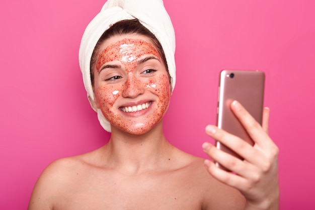 Happy delighted female with soft skin makes selfie while heaving spa procedure, wears white towel, has glad look, poses smiling isolated on pink. People, beauty and skin care concept