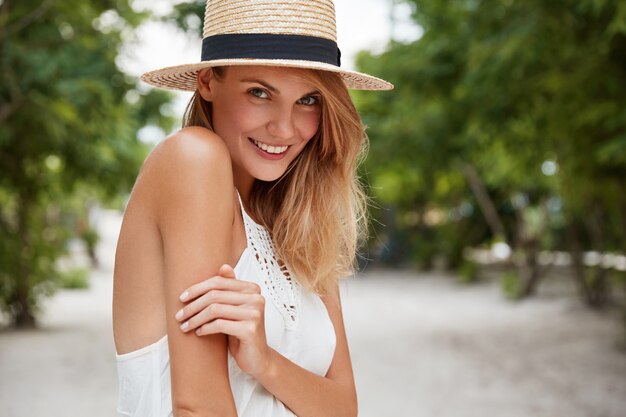 Счастливая, обрадованная женщина имеет приятную привлекательную внешность, носит летнюю шляпу и белое платье, гуляет по улице, любит отпуск или выходной. Довольно молодая зеленоглазая рада женская модель позирует на открытом воздухе